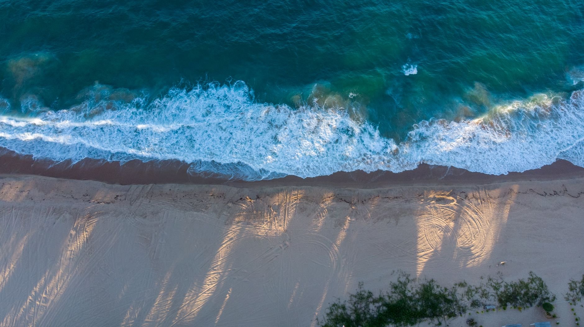 An aerial view of waves breaking on a sandy shore, with long shadows cast across the sand.