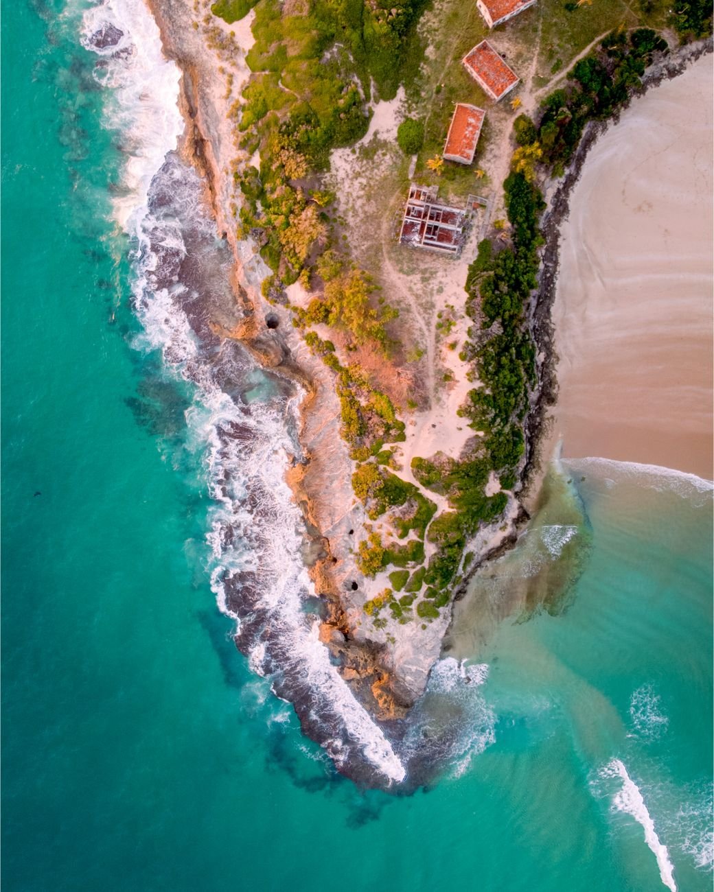An aerial view of a rocky, tree-covered point reaching into turquoise water beside a sandy beach.