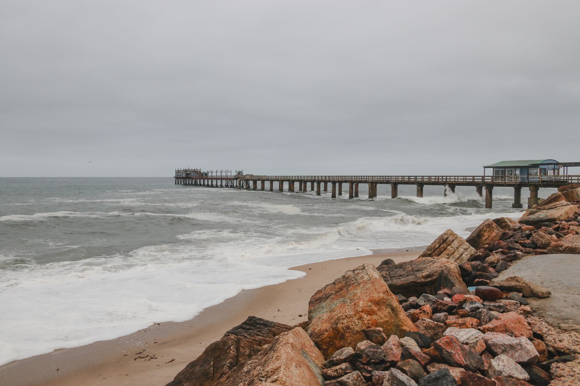 Waves break around the pilings of a long wooden jetty on an overcast day.