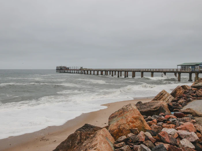Waves break around the pilings of a long wooden jetty on an overcast day.