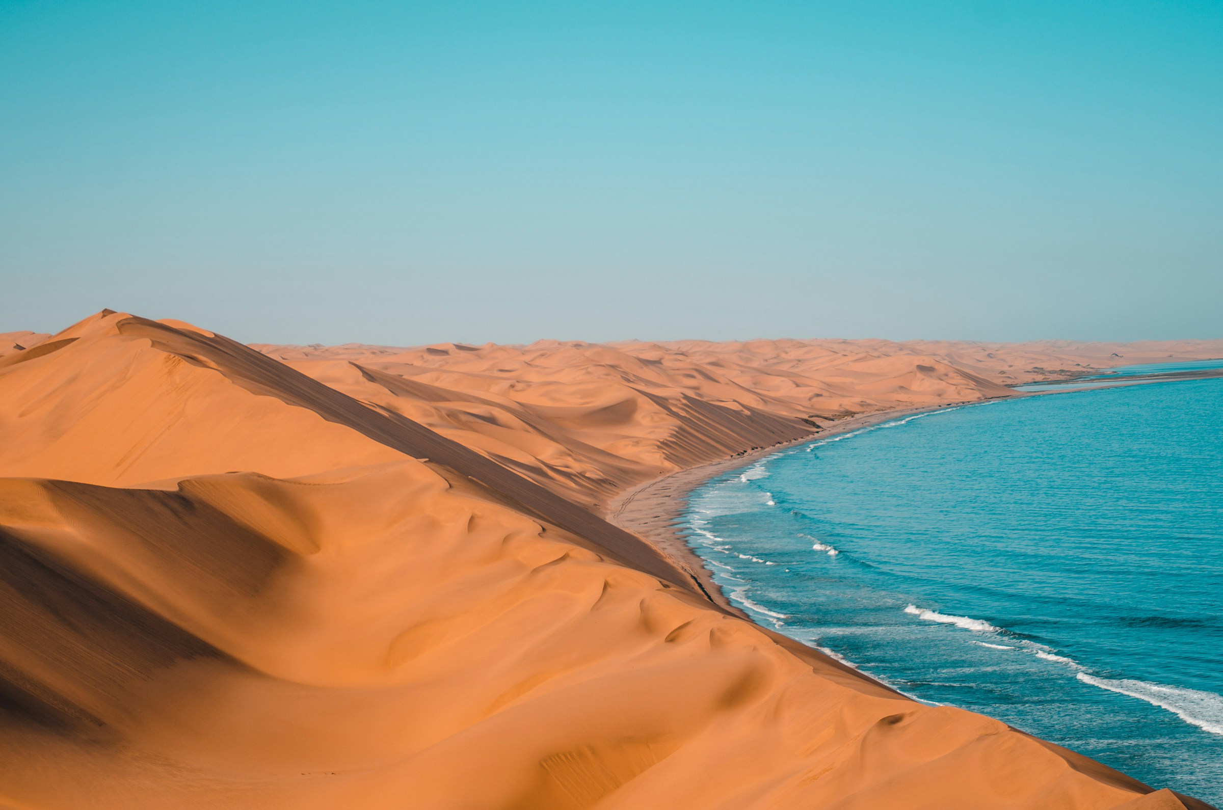A high dune ridge runs down to a curving surf line, with dune fields receding to the horizon and open ocean to the right.