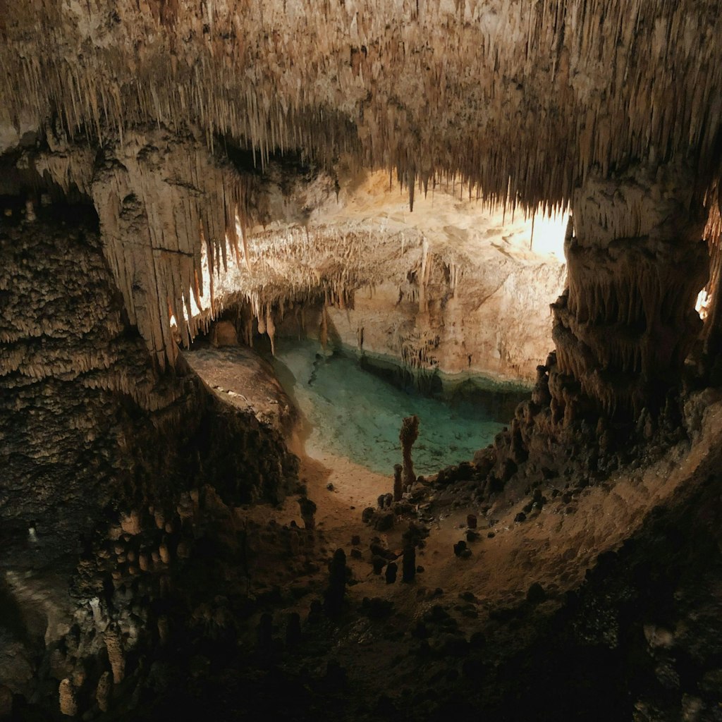 Stalactites and stalagmites surrounding a calm turquoise pool inside a dimly lit cave. Fun Facts About Palma de Mallorca