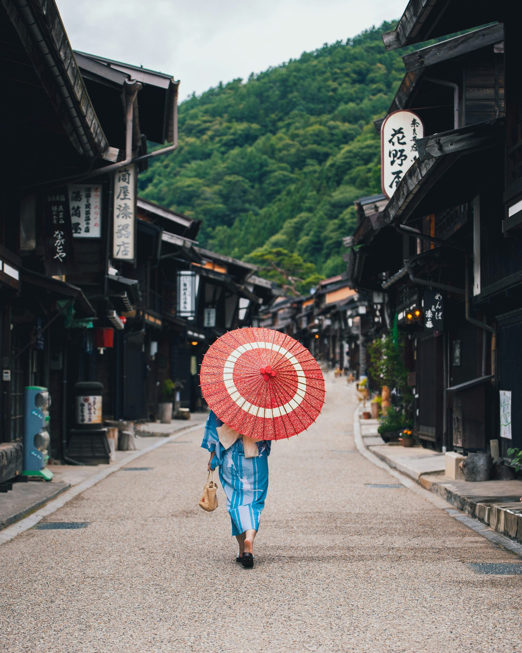 A student in a kimono carries a red umbrella down a traditional street lined with wooden buildings