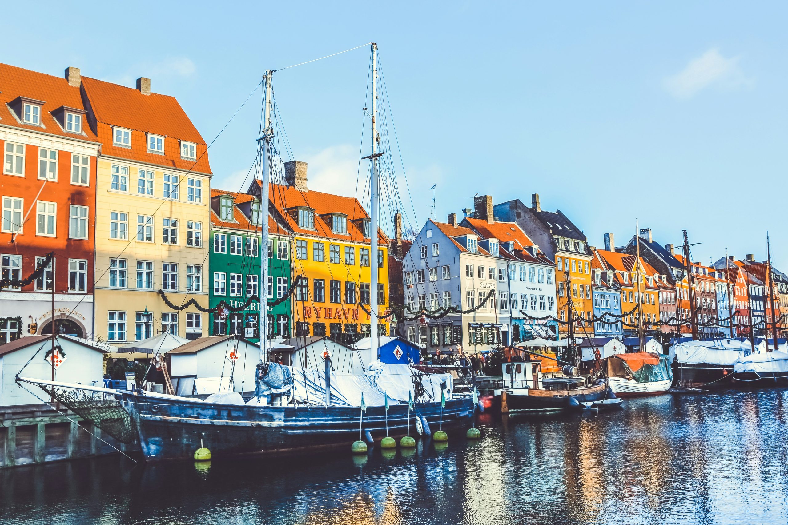 Colourful townhouses line the Nyhavn canal in Copenhagen, wooden sailing boats moored along the quay under a blue sky.