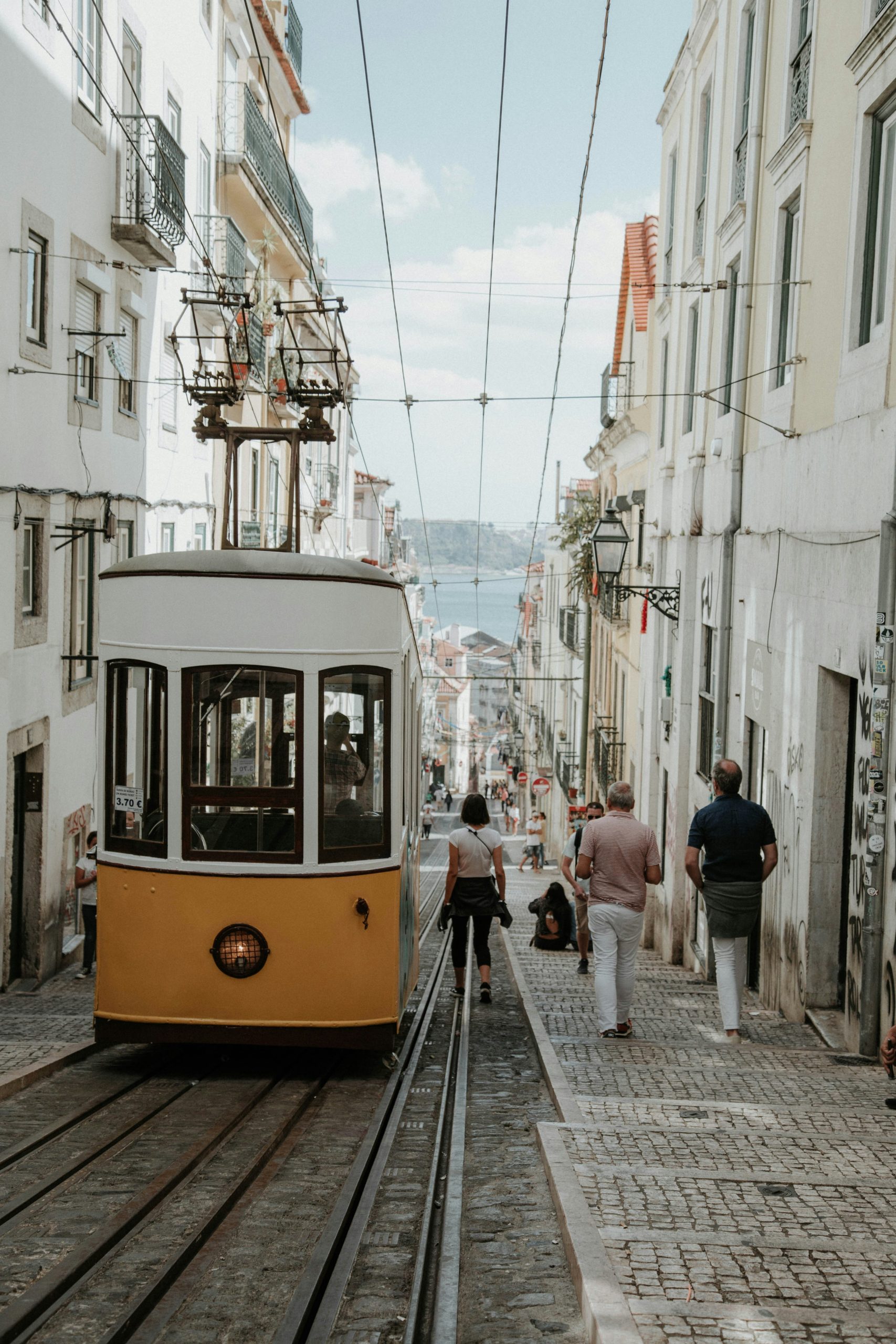 fun facts about portugal. A historic yellow tram moves down a steep, narrow street lined with white buildings. Pedestrians walk alongside the tracks, with a scenic cityscape visible in the background.