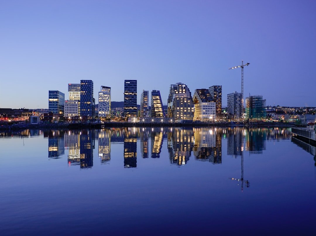 A modern city skyline of angular glass towers glows at dusk, perfectly mirrored in the still harbor water below.