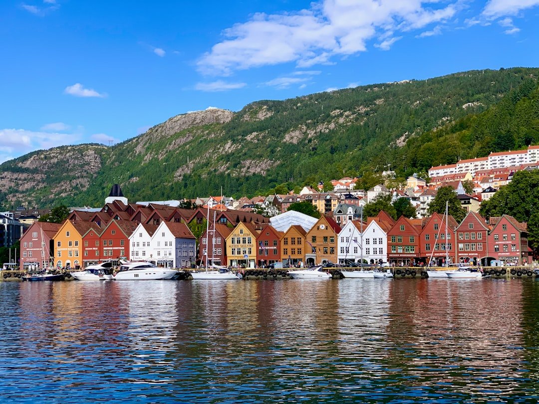Colorful wooden gabled houses line a harbor lined with boats, backed by a forested green mountain under a blue sky.