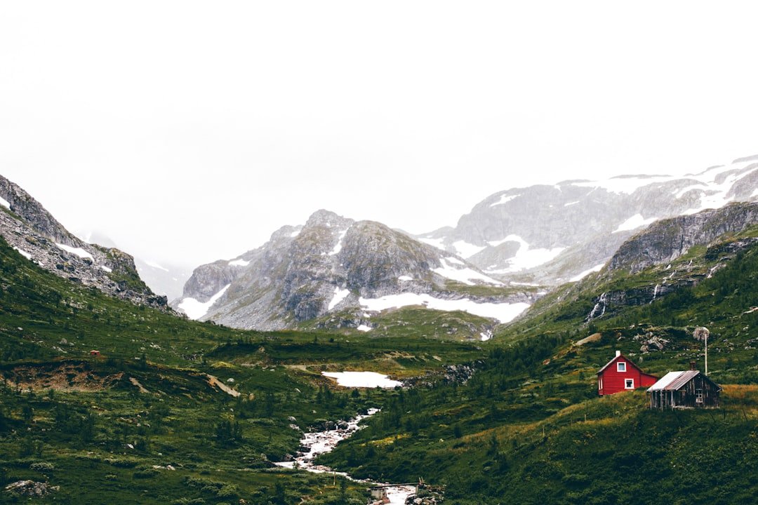 A red timber cabin sits alone in a green mountain valley beneath snow-streaked peaks and low cloud.