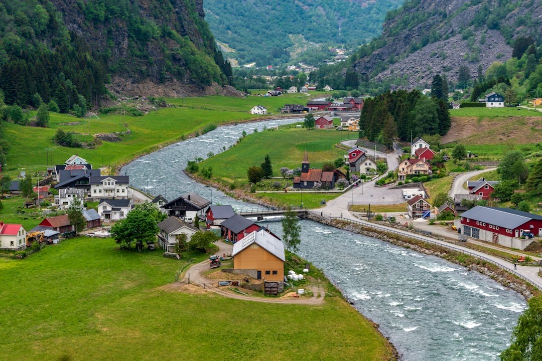 An aerial view of a river valley village with a small white church, scattered houses and green farmland winding between forested mountains.