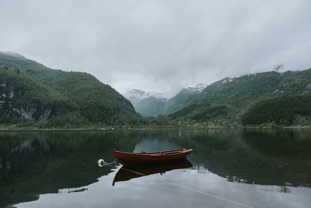An empty wooden rowboat floats on the still, dark water of a fjord surrounded by forested slopes and distant snow-capped peaks.