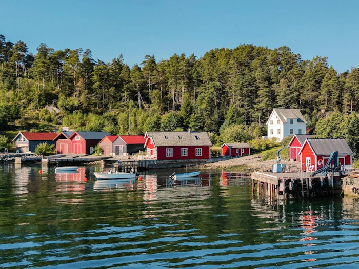 Red and white wooden boathouses sit along a calm fjord shoreline backed by dense pine forest under a clear sky.