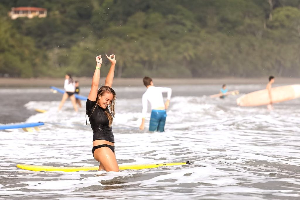 A young woman in a wetsuit raises her arms in excitement after riding a wave on a yellow surfboard, celebrating a victory on an adventure teen tour abroad.