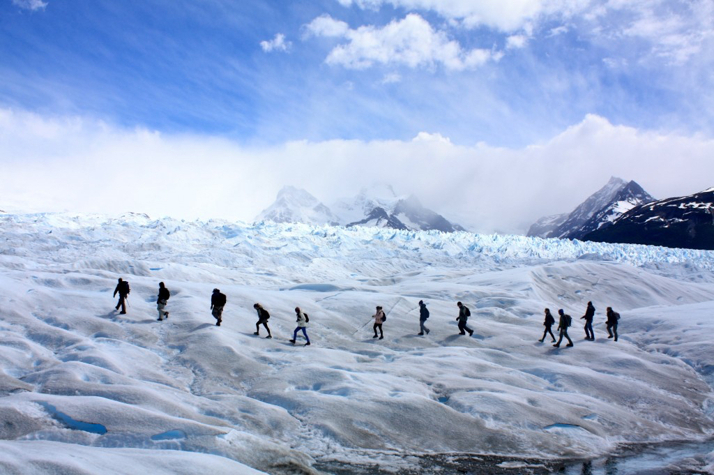 A line of ten walkers crosses a wide field of ridged glacier ice in single file, with dark peaks and low cloud behind them.