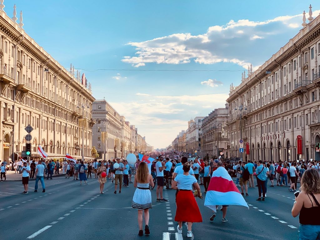 Bilde av Egor Kunovsky: https://www.pexels.com/nb-no/bilde/vei-folkemengde-parade-protest-10994718/ photo of the independence avenue in Minsk, Belarus. People with flags and buildings going down the road