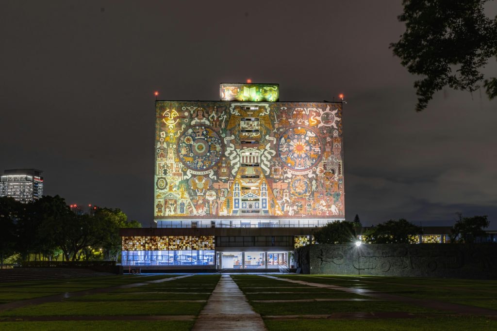 Night view of the Central Library at UNAM in Mexico City, showcasing its illuminated mural filled with historical and cultural symbols.