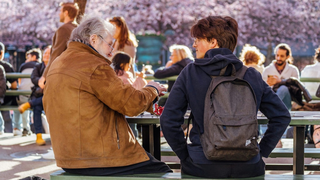 One older man and younger man are playing chess in the park. In the back cherry blossom trees are blooming and people are coming together. #fun facts about Stockholm