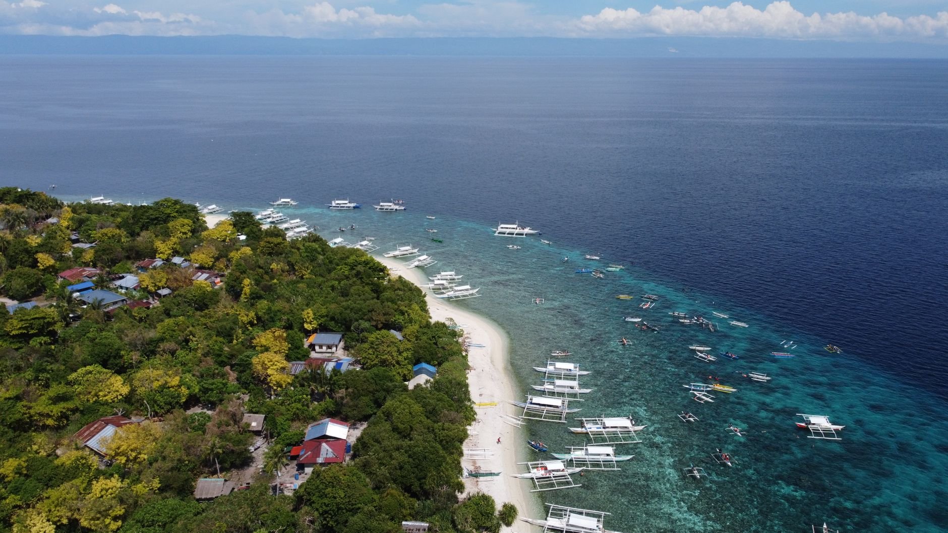 An aerial view of a white sand beach lined with outrigger boats along a turquoise reef.