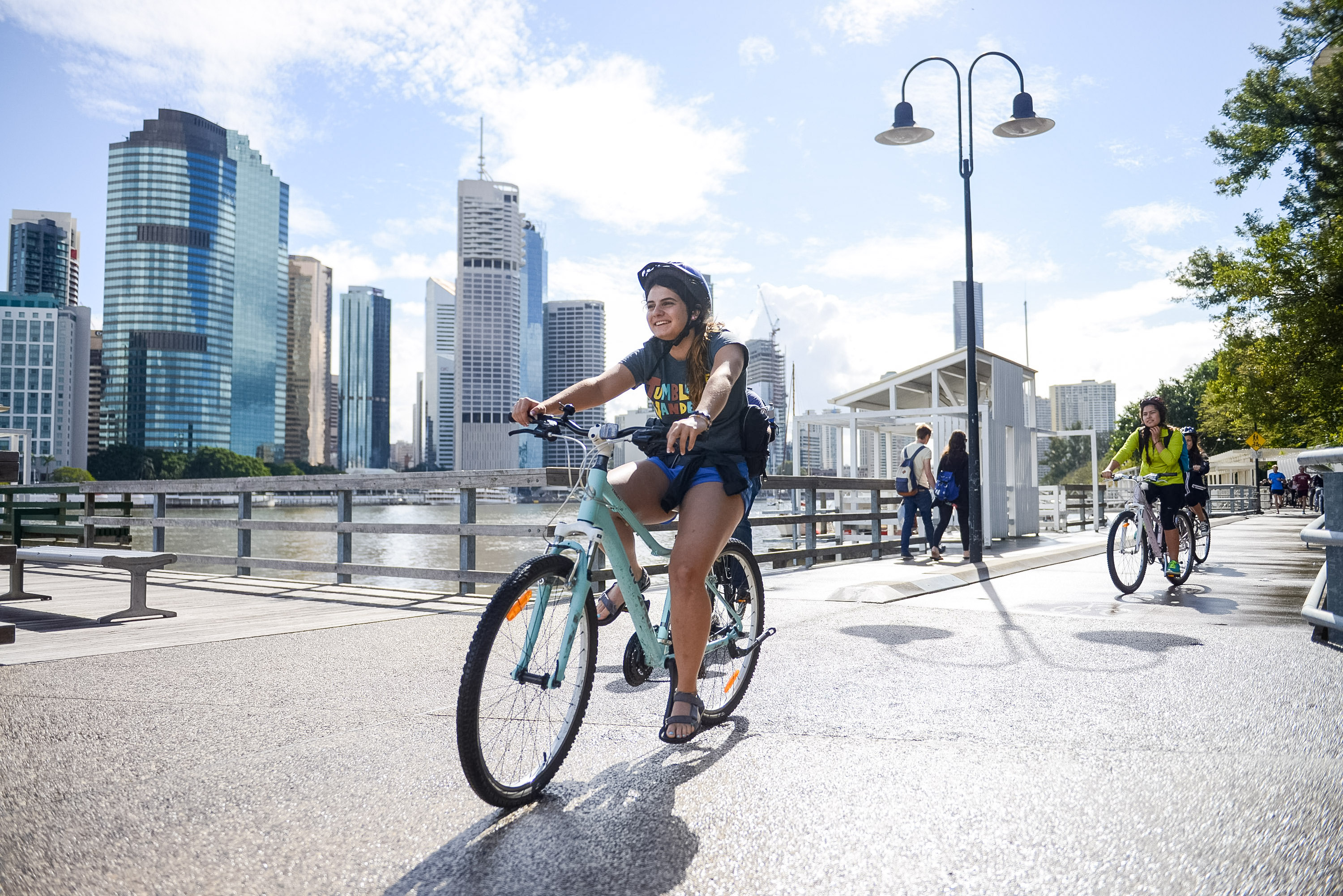 Students ride bikes along a riverside path with a city skyline in the background
