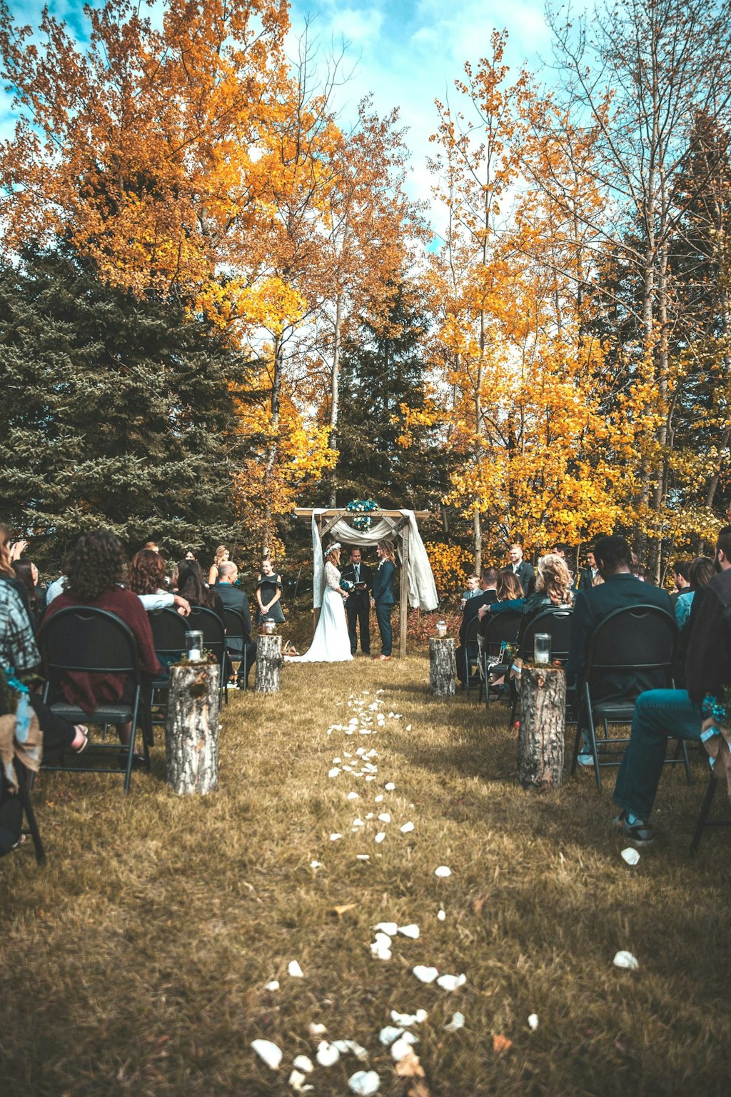 A couple exchanges vows in a garden, surrounded by the colors of autumn. The trees are adorned with leaves in shades of gold, amber, and crimson, creating a warm, natural backdrop. 