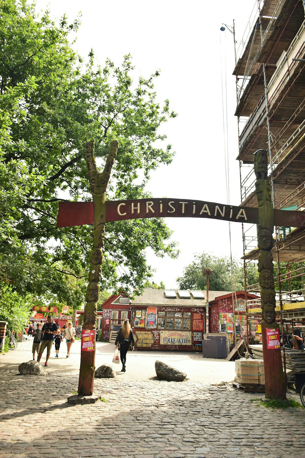 The entrance sign of Christiania (written on the sign) with people, buildings and trees in the back.