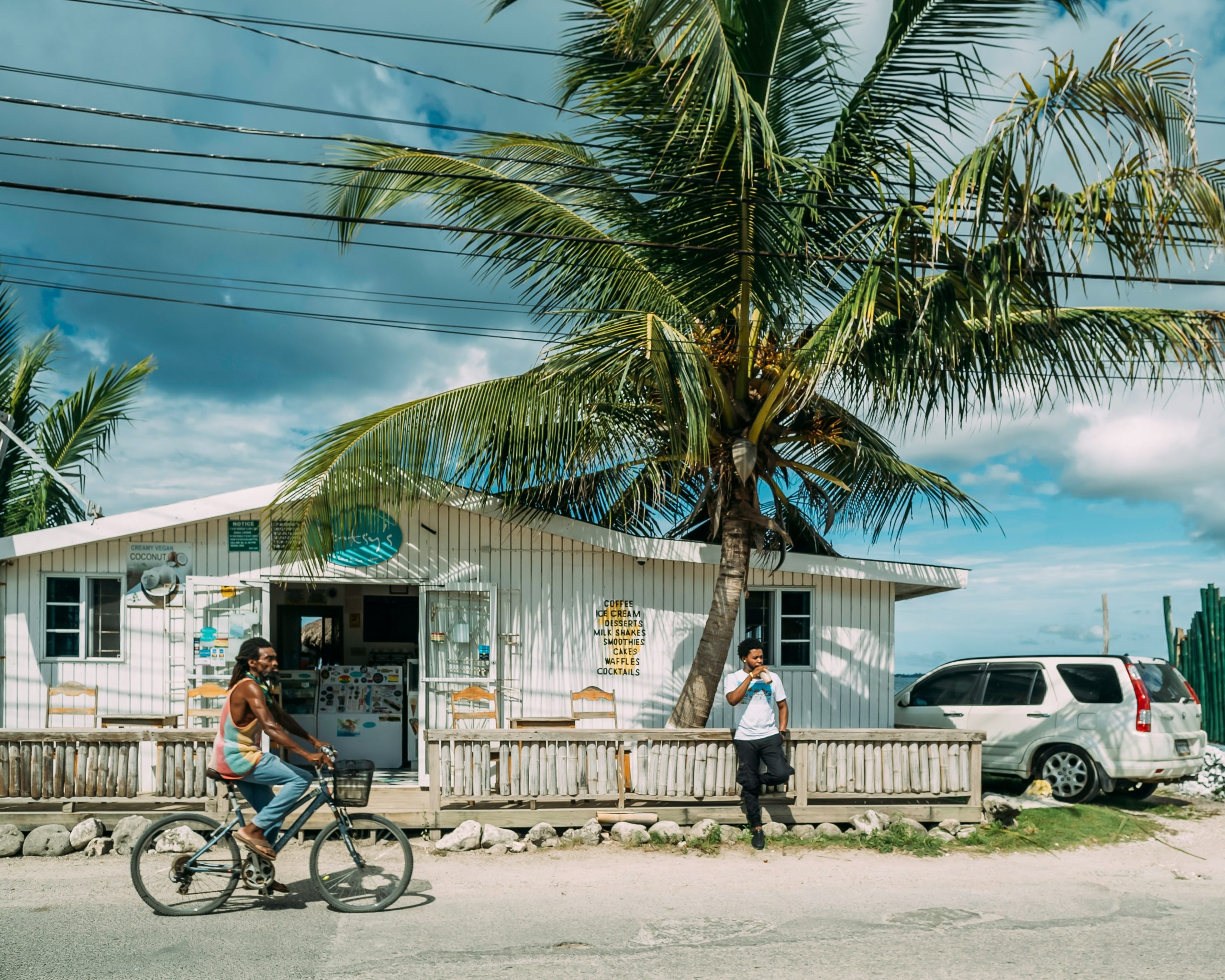 Man riding a bicycle past a small shop with palm trees and another man standing outside in Jamaica.