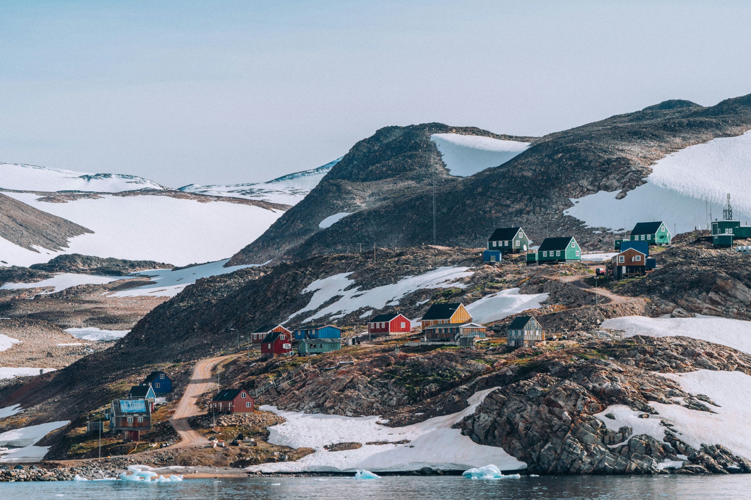 A small village with colorful houses on a snowy and rocky hillside by a lake, with mountains in the background.