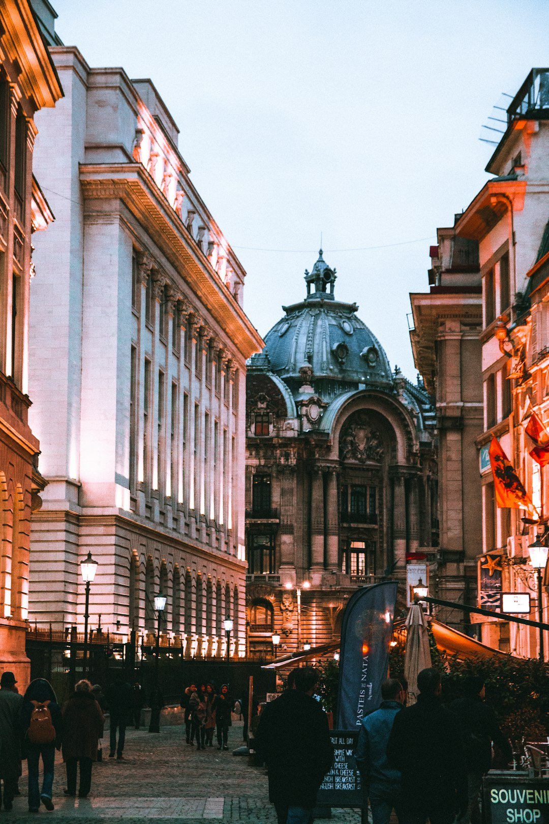 Pedestrians walk along a cobblestone street at dusk beneath illuminated historic facades and a domed building in the distance.