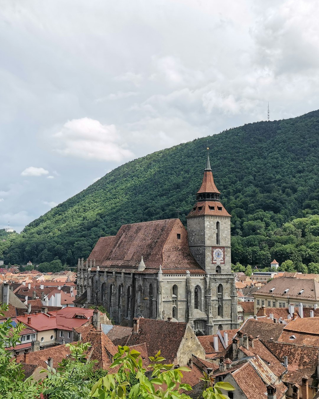 A Gothic church with a tall clock tower and steep red-tiled roof stands among a cluster of orange-roofed houses beneath a forested mountainside.