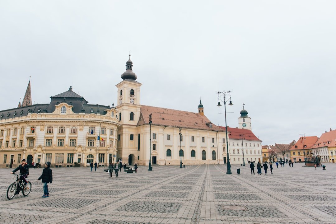 A wide cobblestone square bordered by pastel baroque buildings and a clock tower, with pedestrians and a cyclist crossing in the foreground.