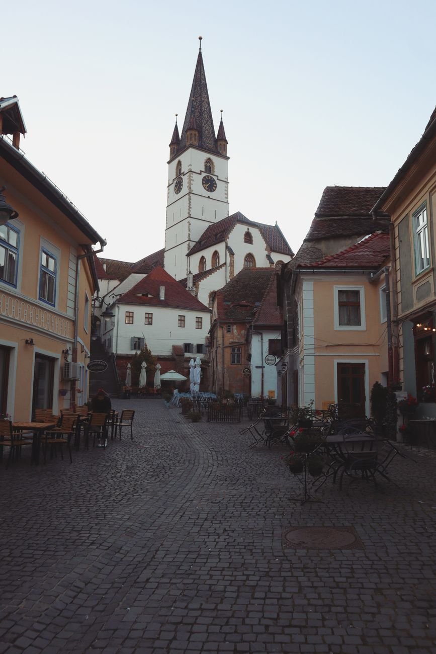 A tall Gothic cathedral spire rises above a row of pastel townhouses along a cobblestone square lined with outdoor cafe tables.