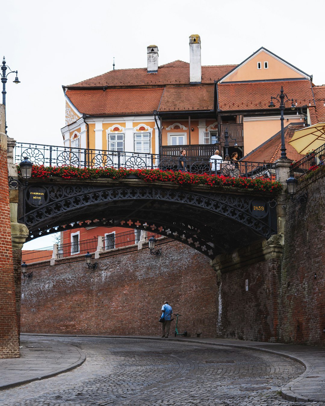 A covered iron pedestrian bridge decorated with red flowers arches over a brick-walled lane between two colorful townhouses.