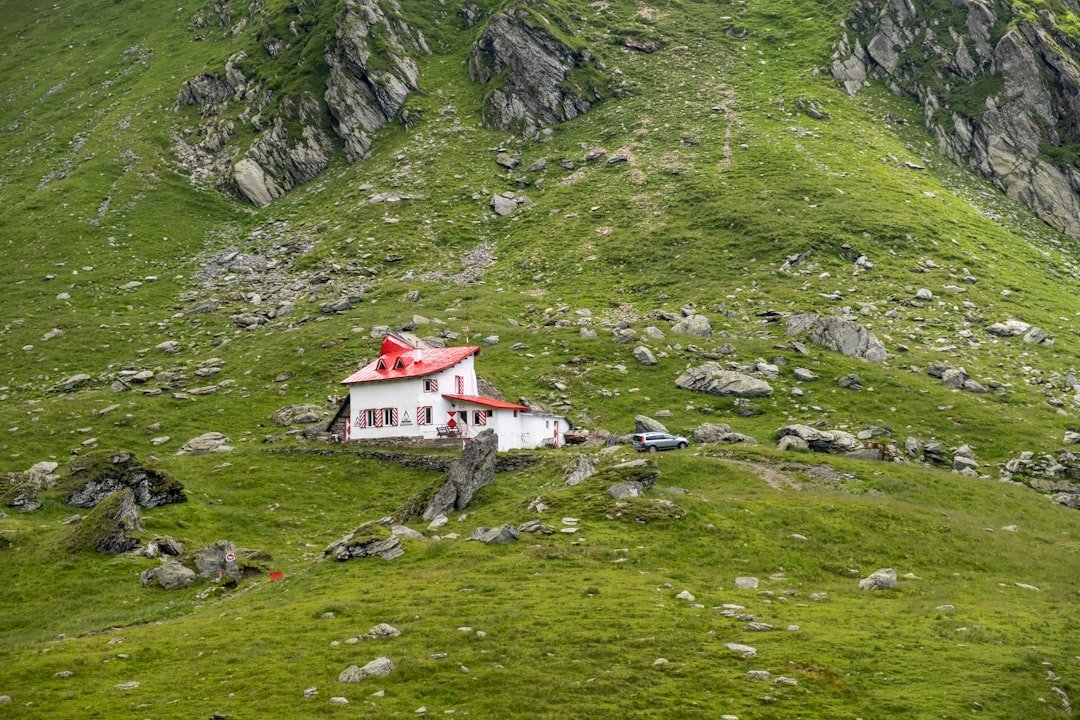 A small white mountain lodge with a red roof sits alone on a steep, rocky alpine slope beside a parked car.