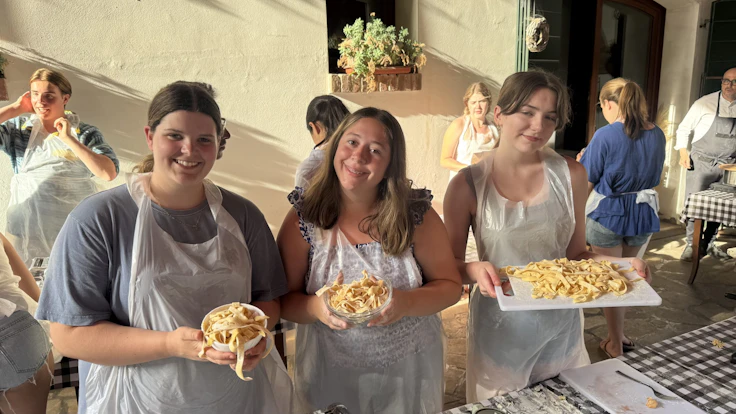 Three teen travelers in aprons hold bowls and a tray of fresh pasta they made in Italy