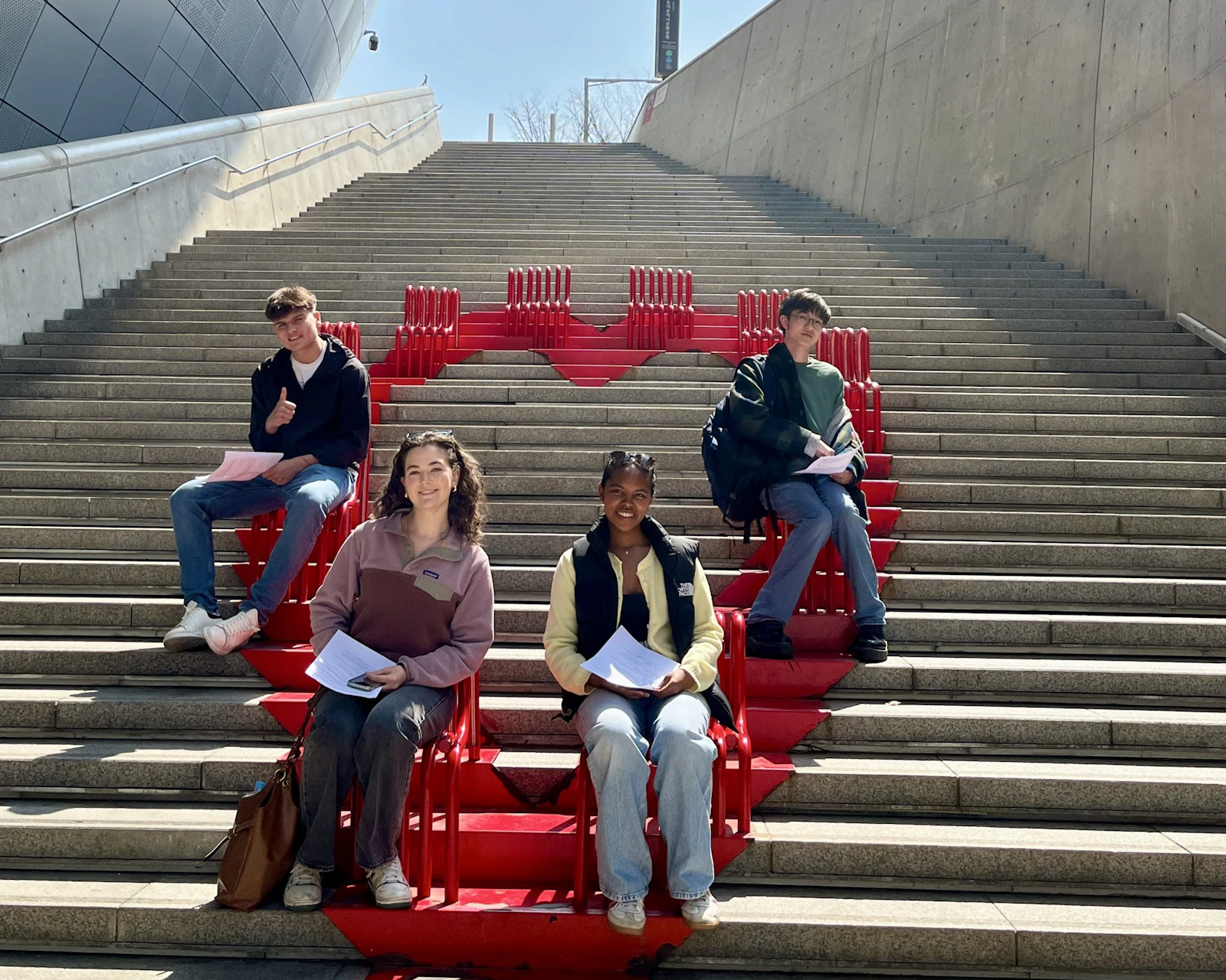 Students sit on red seating built into a long outdoor staircase at Dongdaemun Design Plaza in Seoul