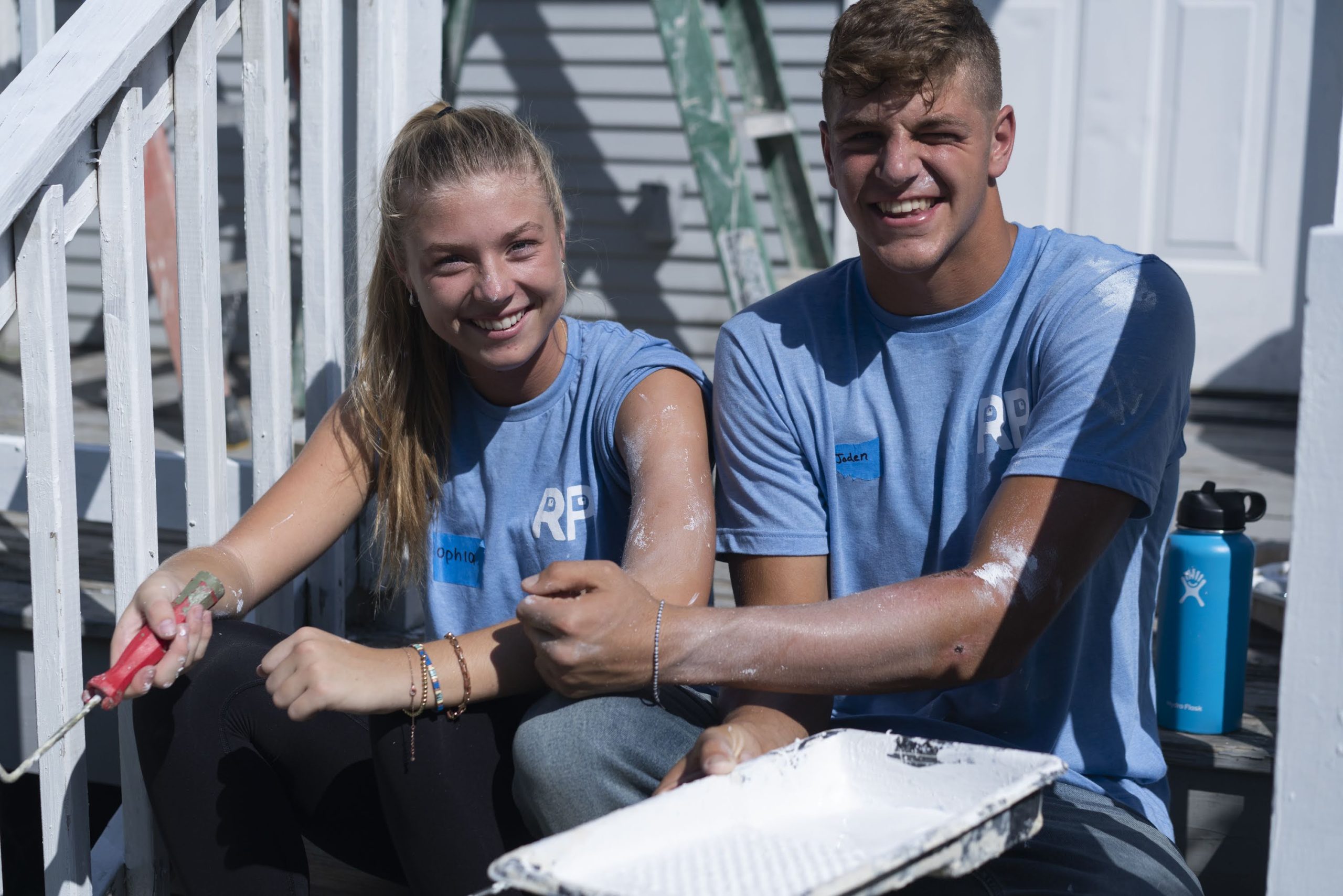 A smiling young woman and man, both wearing casual summer clothes, sit down outside of a house with painting gear.