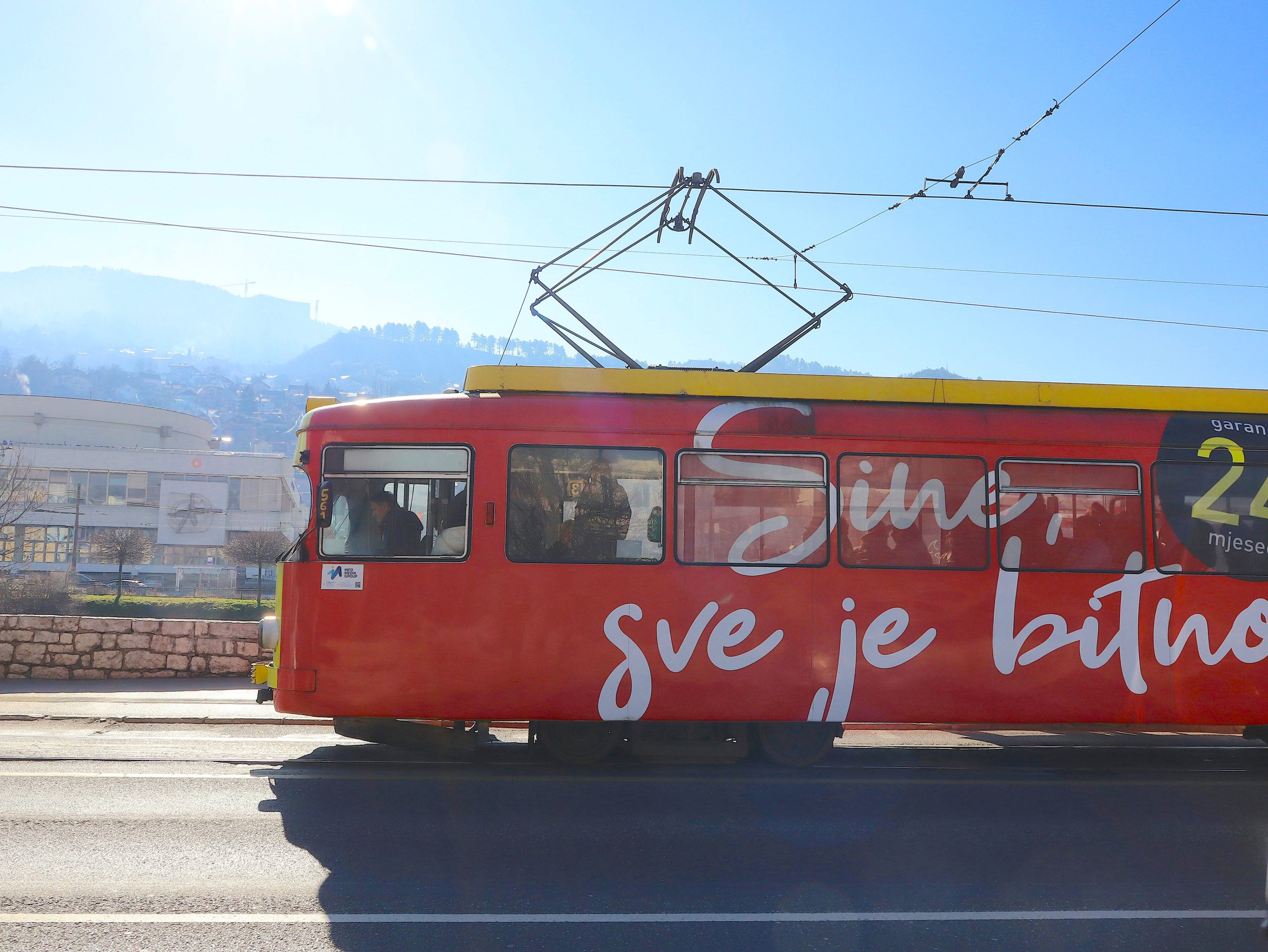A red and yellow tram with white script lettering along its side runs on a city street under a bright sky, hills behind.
