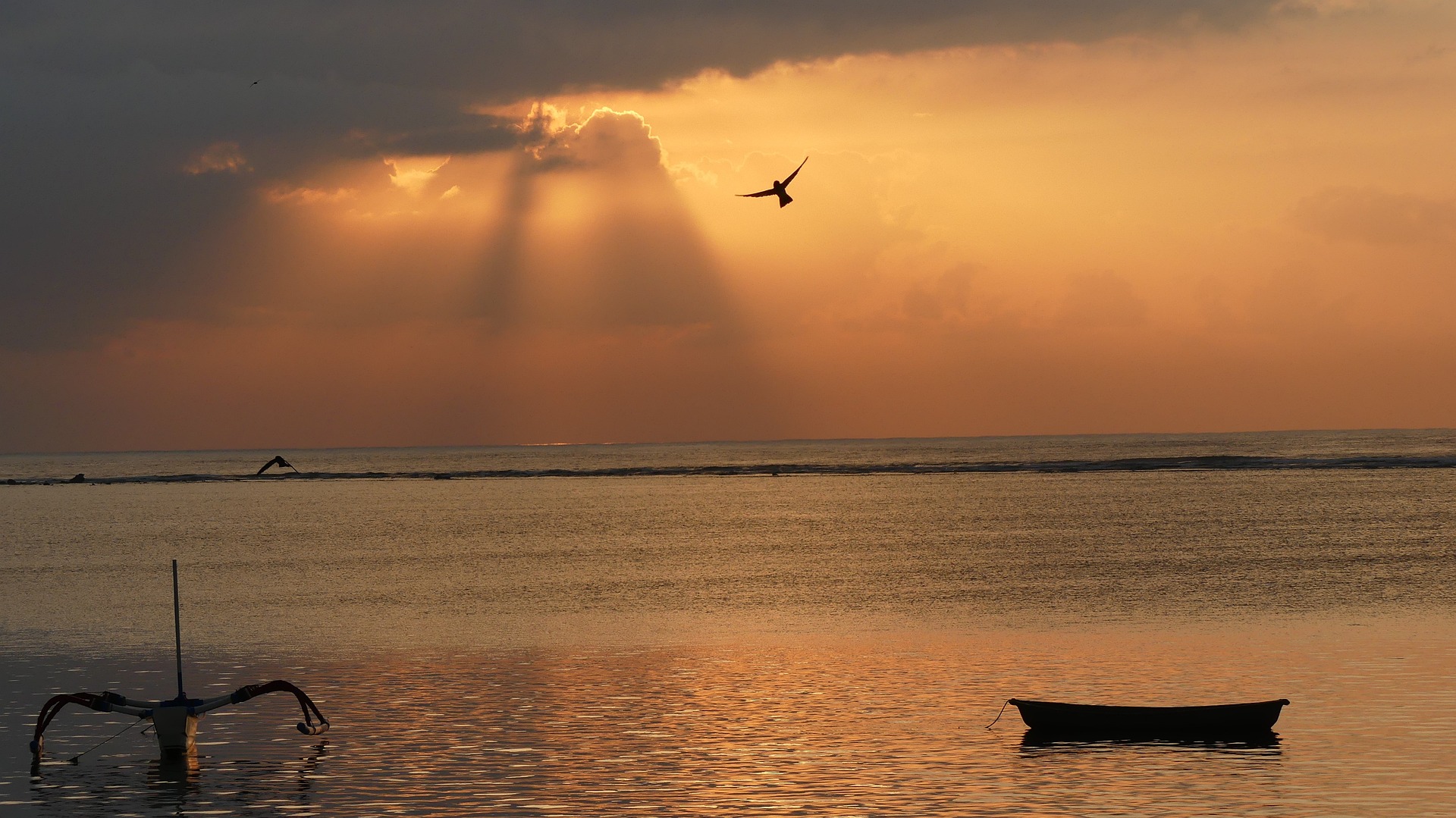 Small boat floats offshore under an orange sunset sky with a bird in flight
