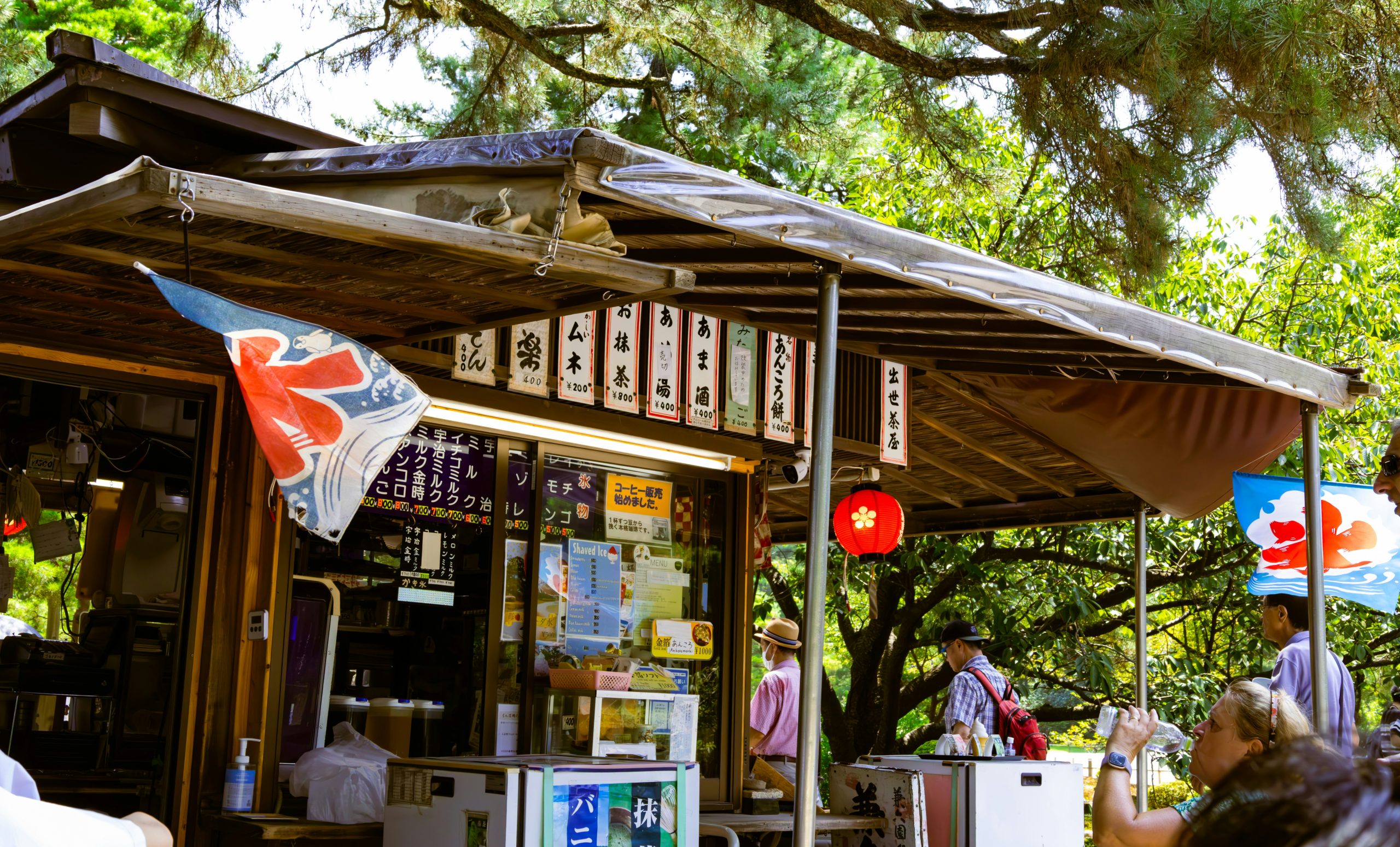A traditional Japanese food stall with hanging signs sits beneath trees as visitors gather nearby