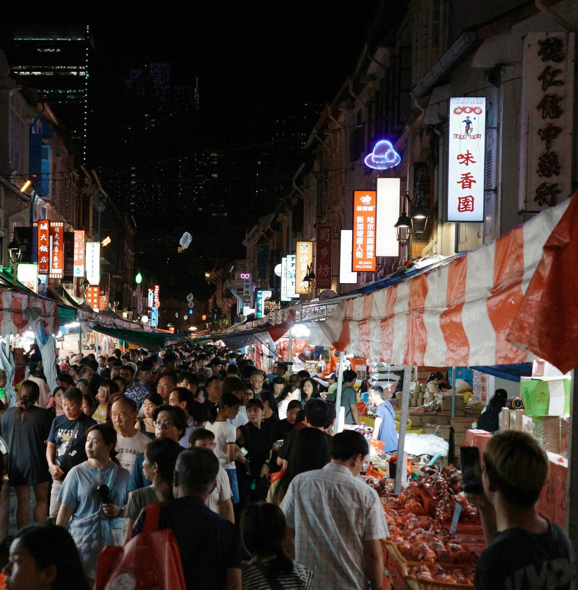 A crowded night market in Chinatown, stalls under red and white striped awnings on both sides of a narrow street, lit shop signs in Chinese above them and office towers behind.
