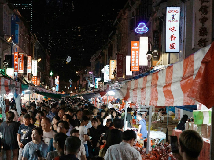 A crowded night market in Chinatown, stalls under red and white striped awnings on both sides of a narrow street, lit shop signs in Chinese above them and office towers behind.