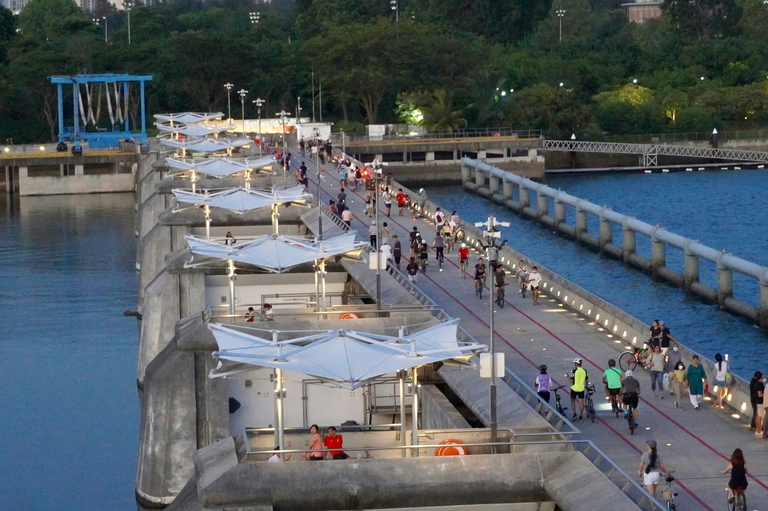 The Marina Barrage dam at dusk, its long deck lit and lined with white canopies, people walking and cycling along it with the reservoir on one side and the sea on the other.