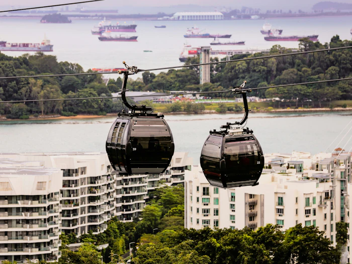 Two cable cars hang from their line above apartment blocks and trees on the way to Sentosa, with container ships at anchor in the harbour beyond.