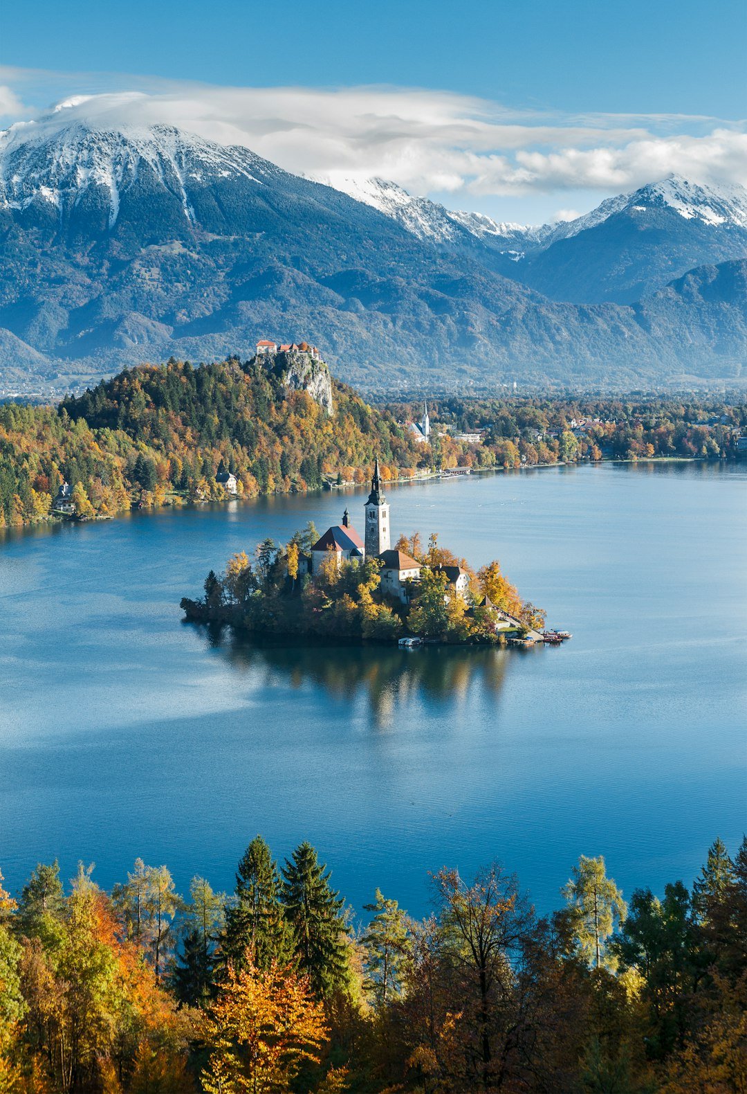 A small island with a church and bell tower surrounded by still water, trees, and mountains.