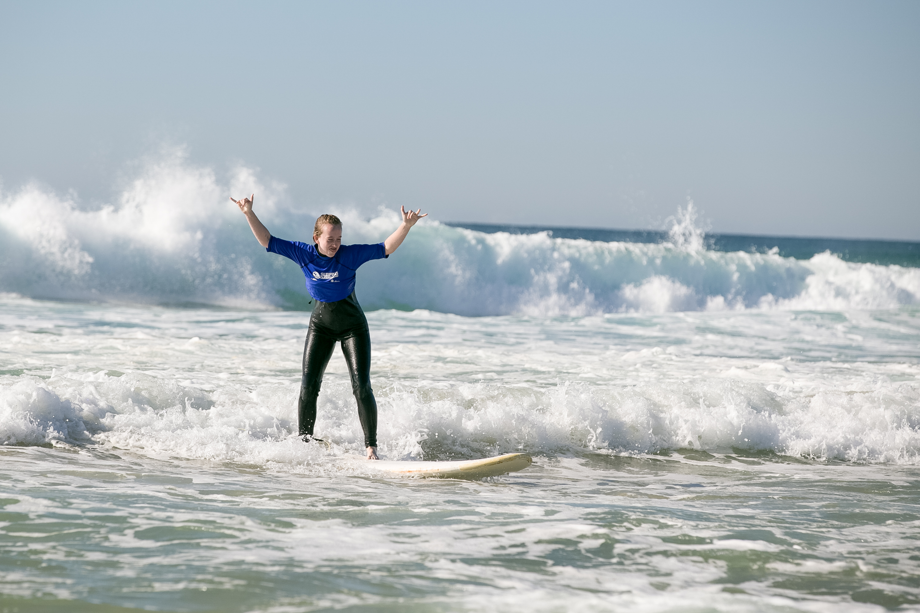 A student throws up their arms while riding a surfboard through breaking waves