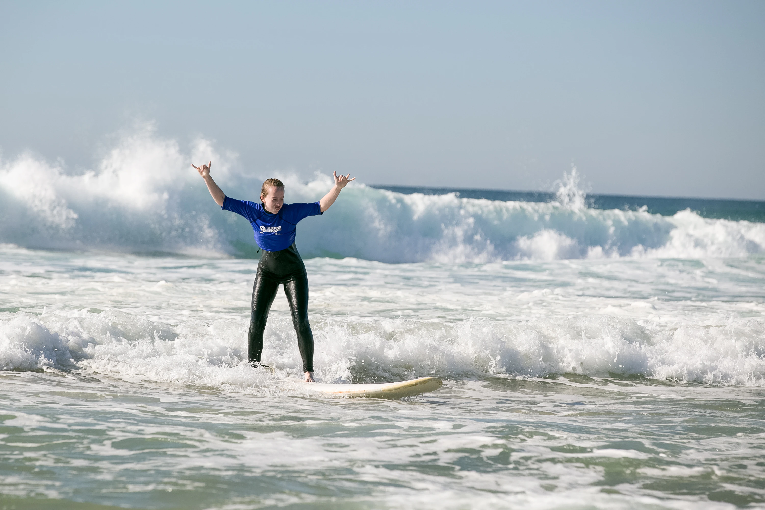 A student throws up their arms while riding a surfboard through breaking waves