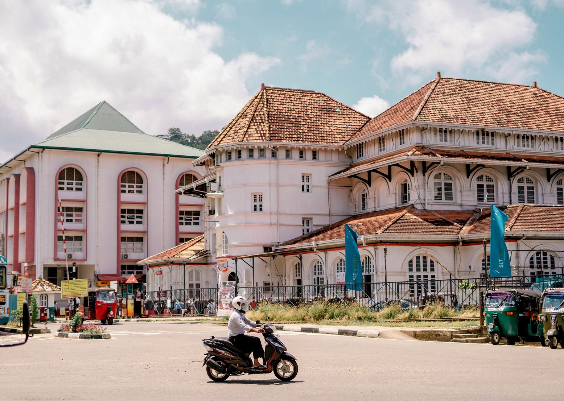 A scooter passes shopfronts on a street beside a post office.