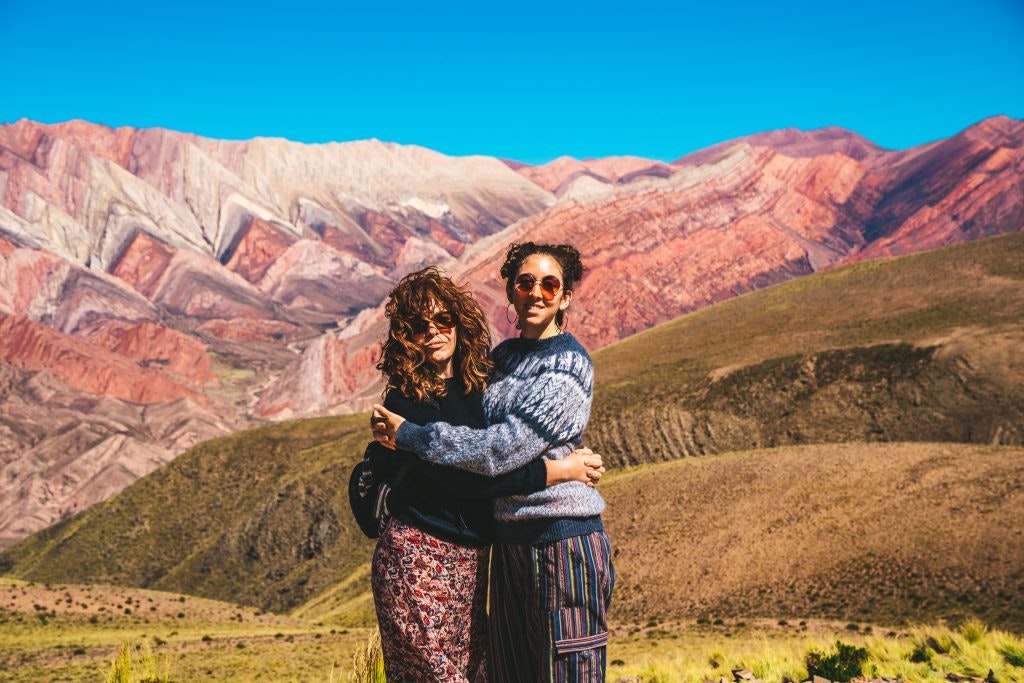 A medium shot of two people embracing and smiling on a grassy hill, with the dramatic, multi-colored peaks of the Serranía de Hornocal (Mountain of 14 Colors) in Jujuy, Argentina, sweeping across the background under a clear blue sky. The mountain range features sharp, zigzagging rock formations layered in striking shades of red, pink, ochre, and white, while a sunlit, rolling green and brown ridge sits in the middle ground.