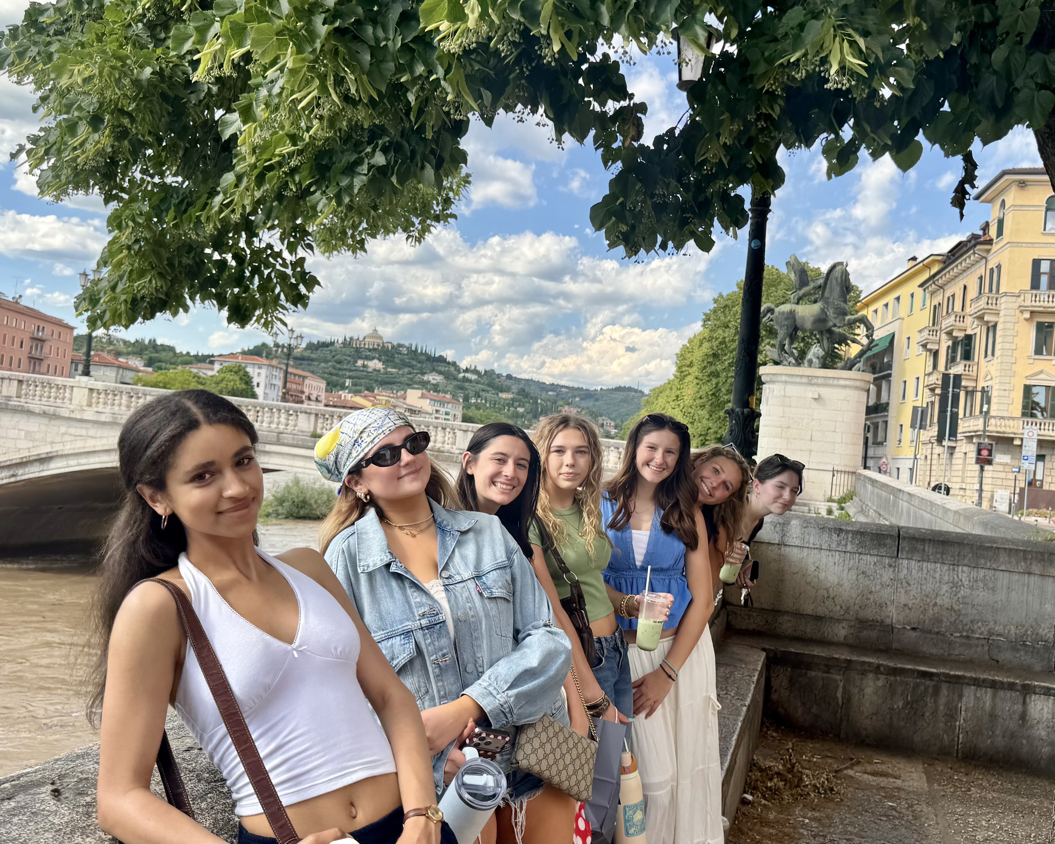 High school students on a Rustic Pathways Italy program posing along the Adige River in Verona, with a historic stone bridge and tree-covered hillsides behind them