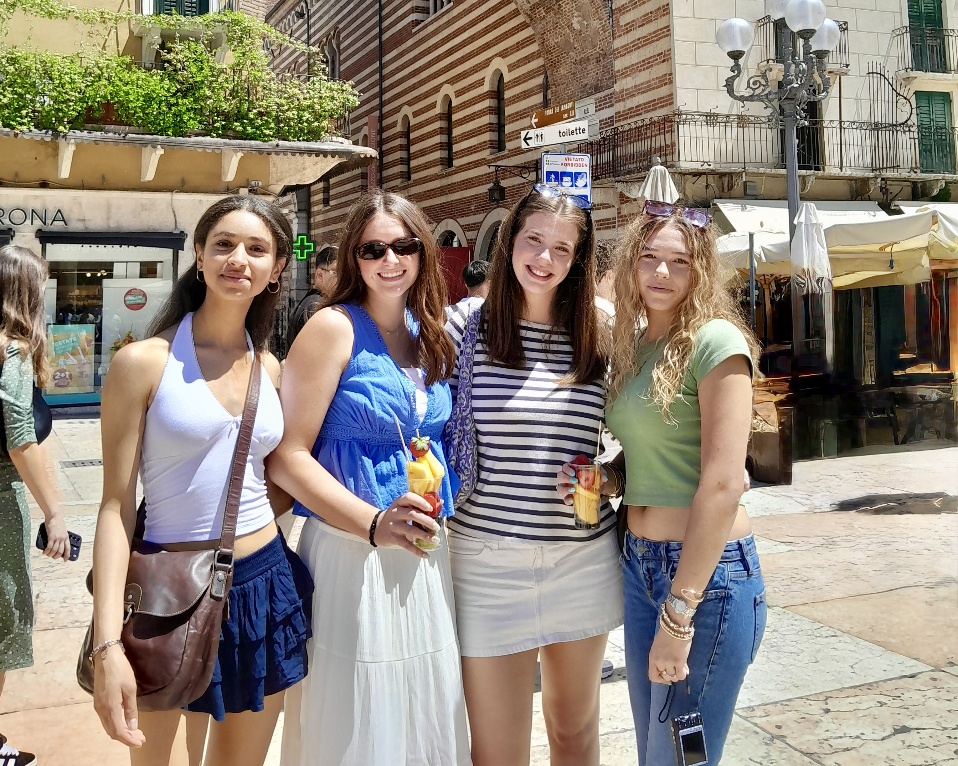 Four high school students enjoying fresh fruit drinks in a sunny Verona piazza, with the city's striped Romanesque buildings behind them, in Italy