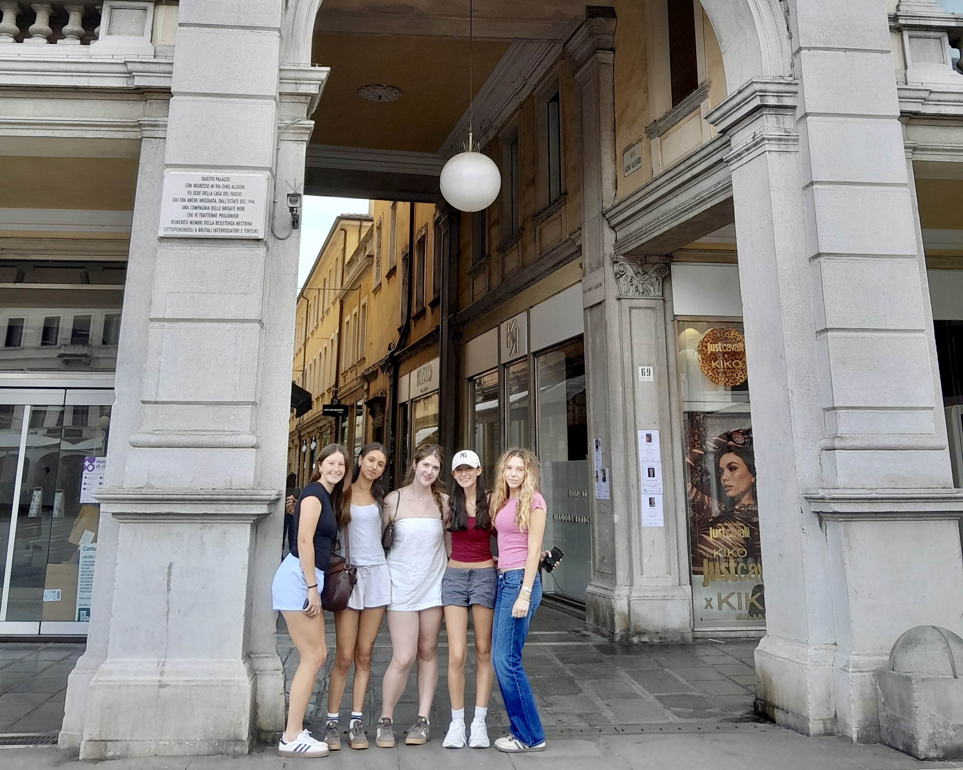 Five high school students on a Rustic Pathways Italy program posing under a white marble portico arch on a pedestrian street in Verona's historic center, Italy